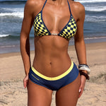 Model wearing a navy blue bikini with a yellow and navy checkerboard pattern on the top and a solid yellow waistband on the bottom, standing on a sandy beach with the ocean in the background.