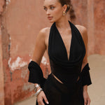 Model wearing a black halter neck dress standing in a outdoor setting with a beige background 