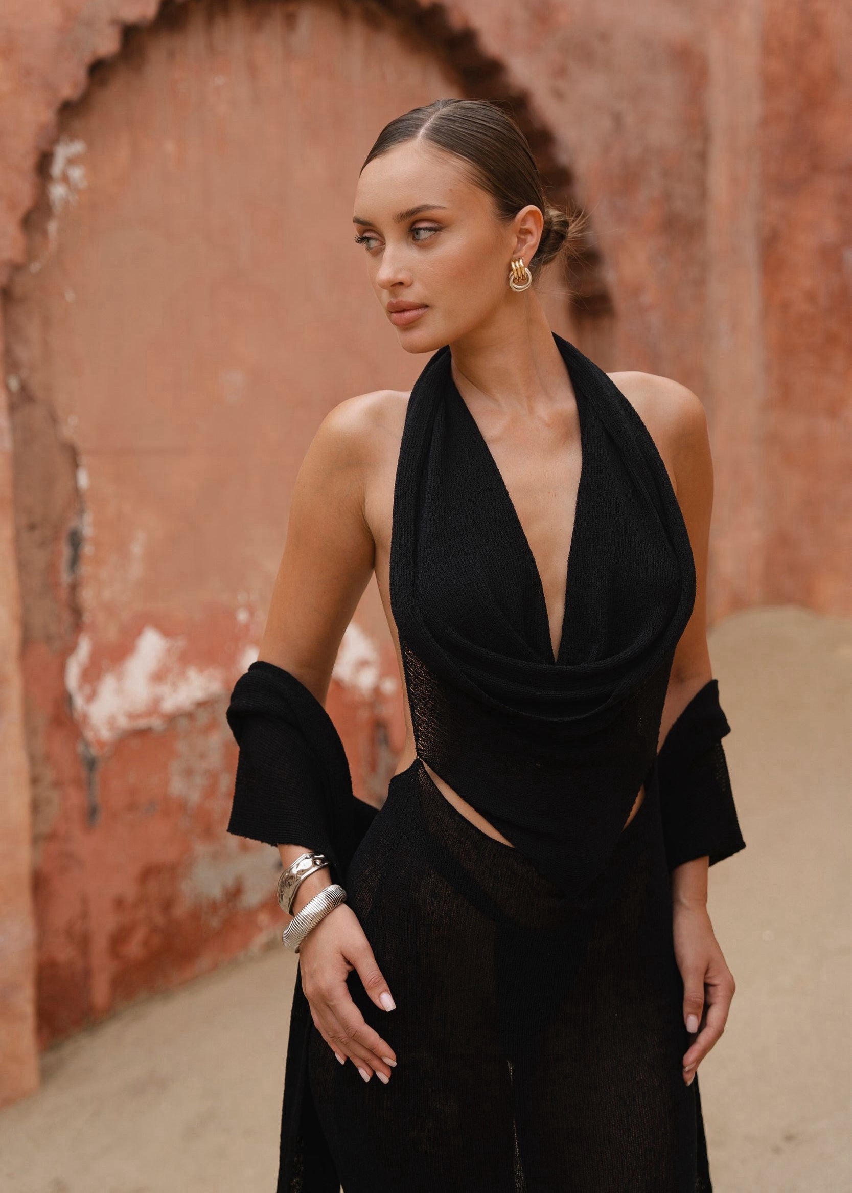 Model wearing a black halter neck dress standing in a outdoor setting with a beige background 