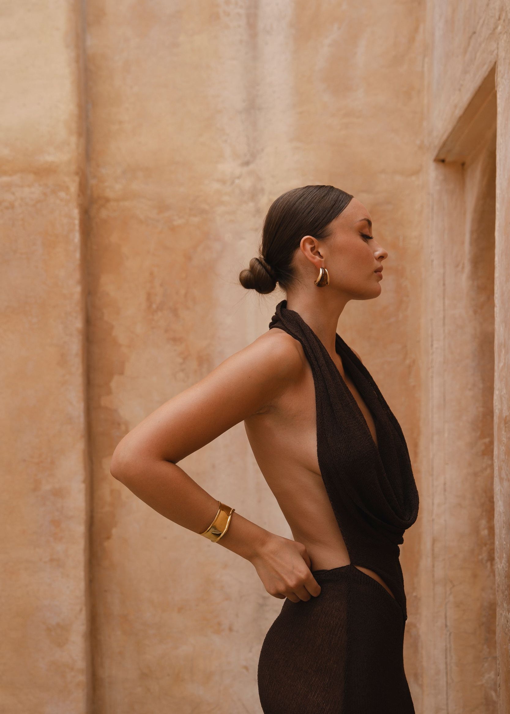 Side shot of model wearing a brown halter neck maxi dress with sand-coloured walls in the background.
