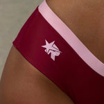 Close-up of a model wearing low-rise bikini bottoms in cherry with a light pink waistband and a pink stylized star logo on the side.  