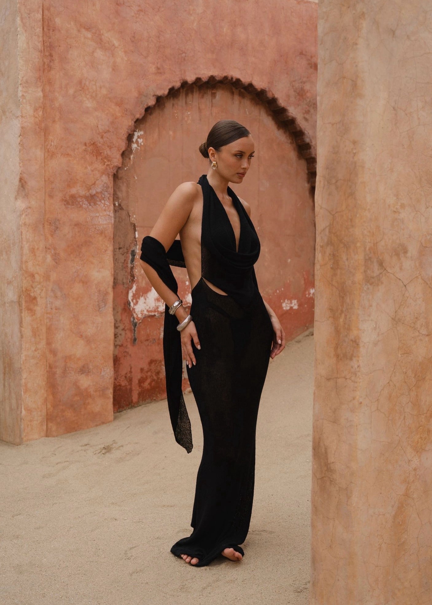 Model wearing a black halter neck maxi dress standing in a sunlit archway with sand-coloured walls.