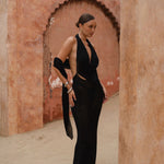 Model wearing a black halter neck maxi dress standing in a sunlit archway with sand-coloured walls.