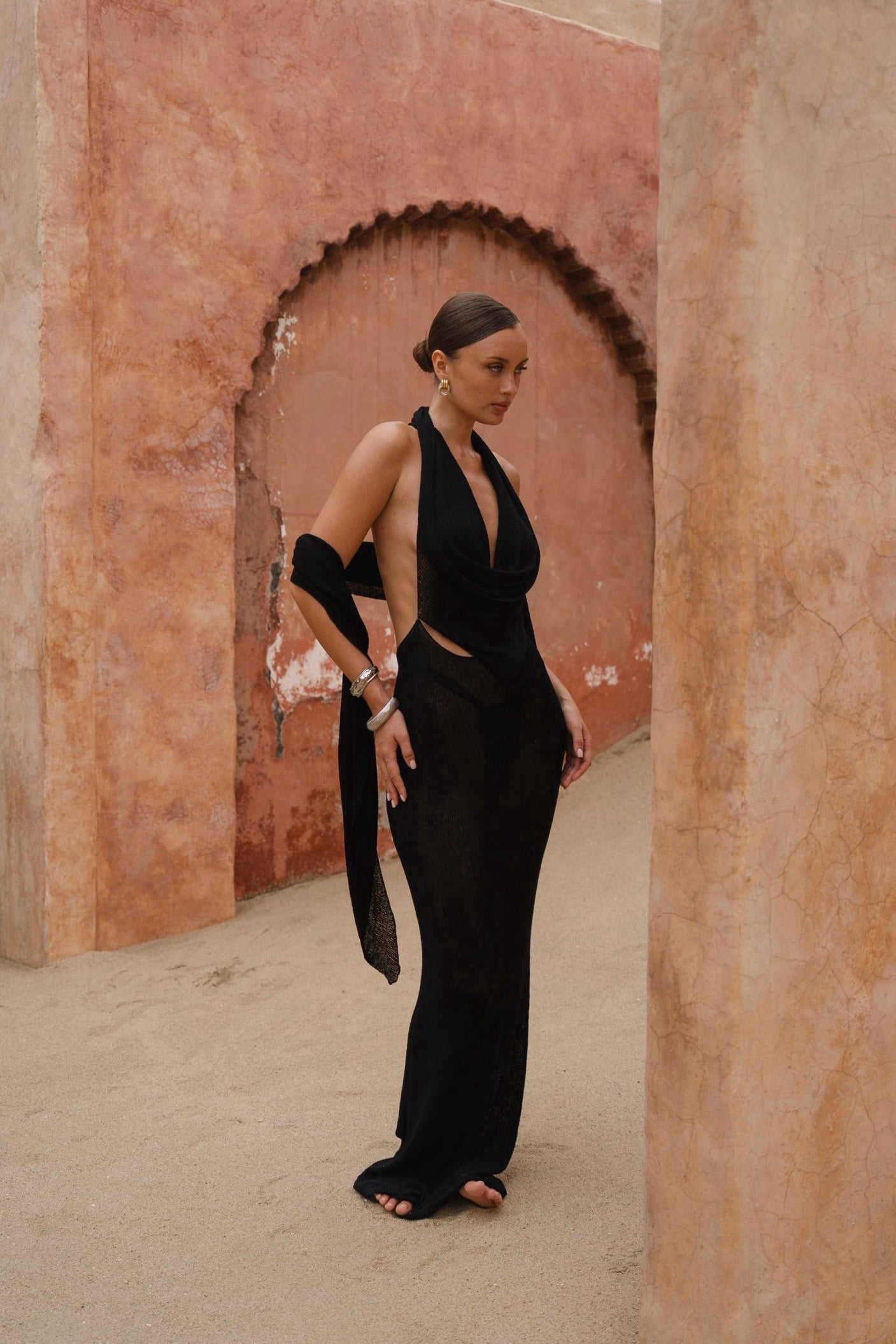 Model wearing a black halter neck maxi dress standing in a sunlit archway with sand-coloured walls.
