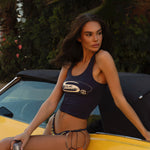 Model wearing a navy Rogue Season graphic tank top with yellow logo, and navy and yellow checkered bikini bottoms, photographed outdoors leaning on a yellow car.