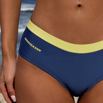 Close-up of model wearing navy blue bikini bottoms with a yellow waistband and yellow 'ROGUESEASON' text on the side, photographed outdoors on a sandy beach.