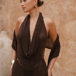 Close up shot of model wearing a brown halter neck maxi dress standing in a sunlit outdoor setting with sand-coloured walls.