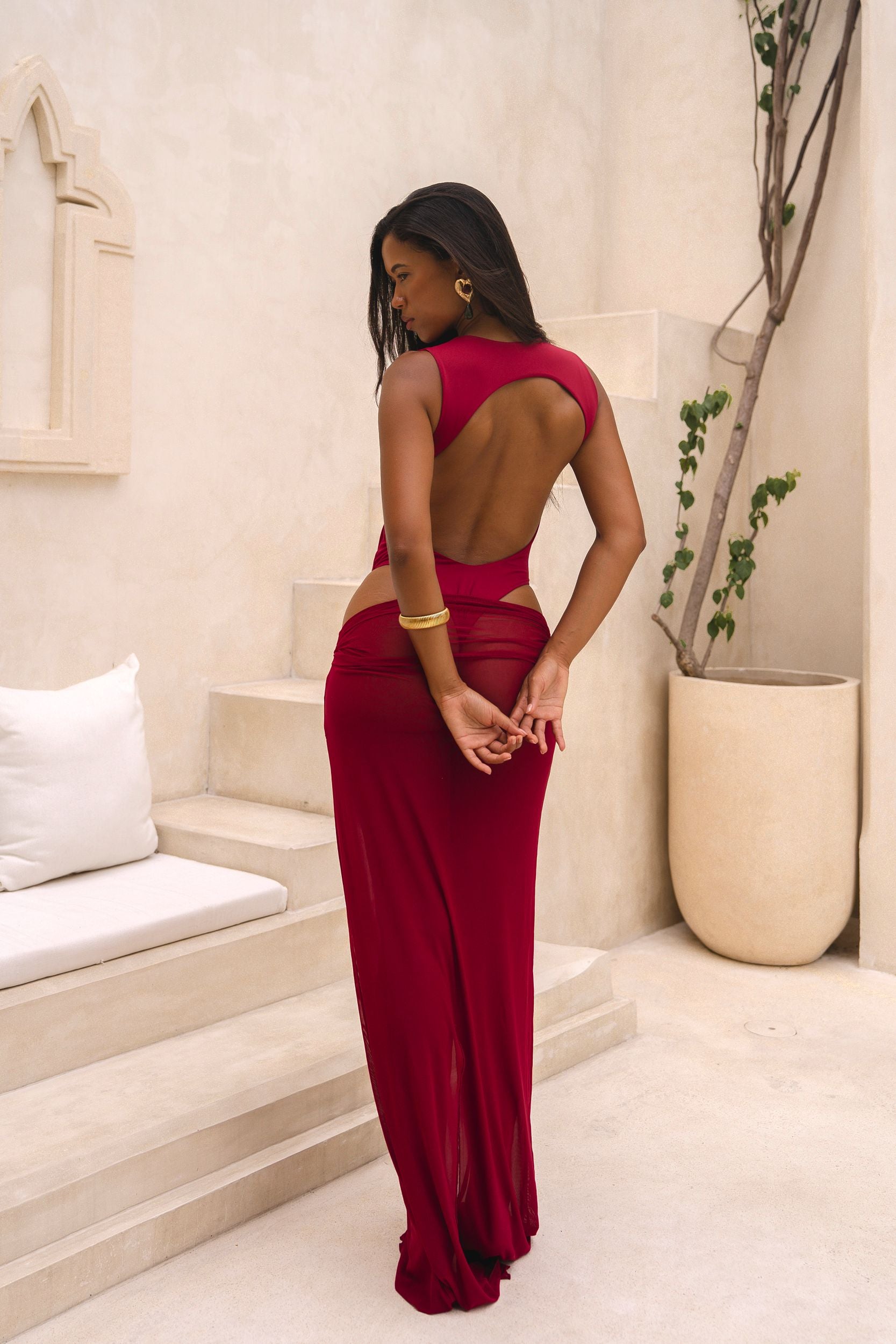Back of model wearing a cherry Rogue Season one-piece swimsuit with side cut-out and matching sheer skirt, standing indoors in a neutral setting.