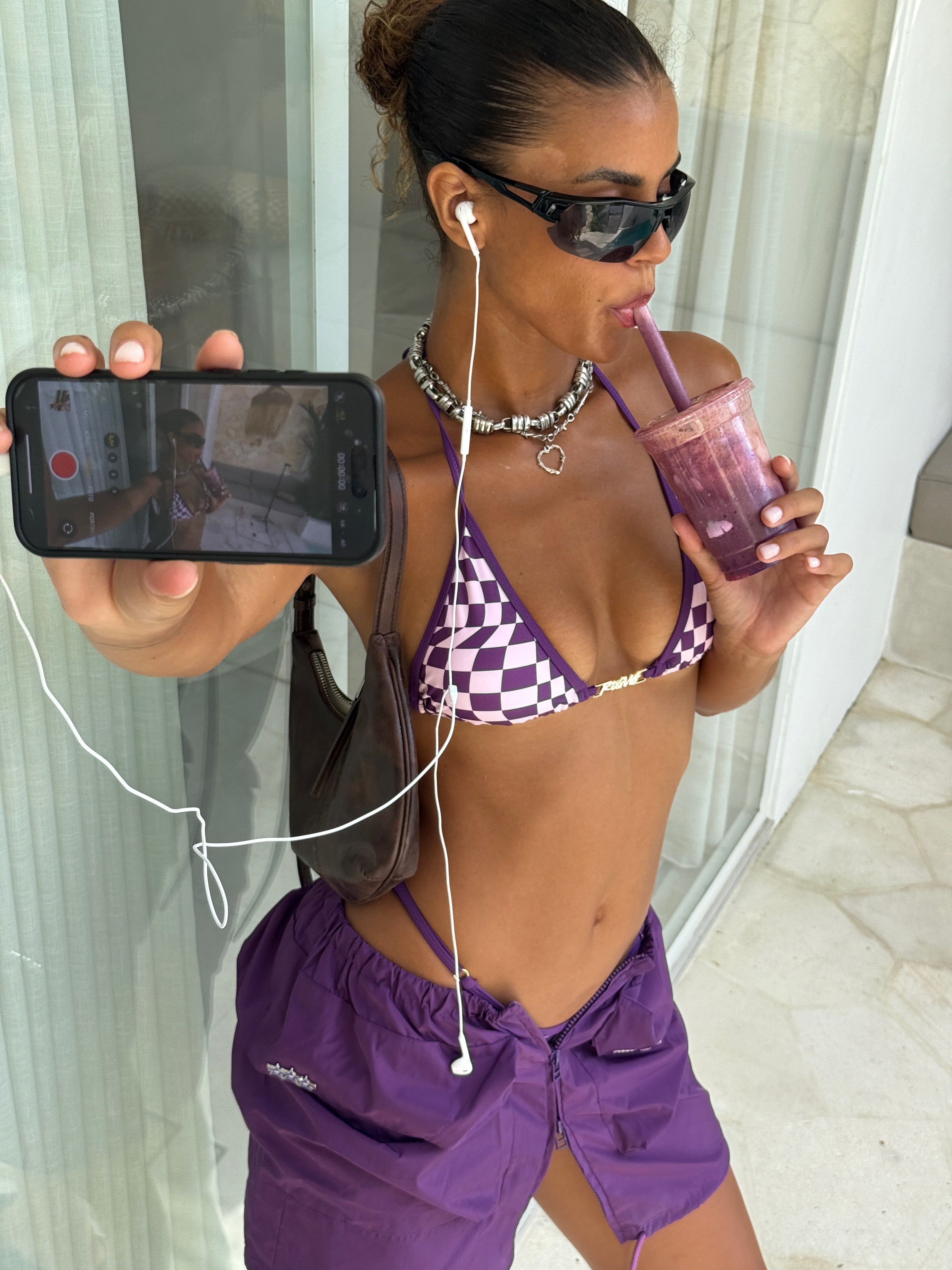 Model in a purple checkered bikini top and matching skirt taking a selfie with a phone, drinking from a smoothie cup.