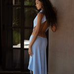 Model wearing a light blue sleeveless backless top with a matching skirt, stands in profile against a beige wall next to a dark-framed window.