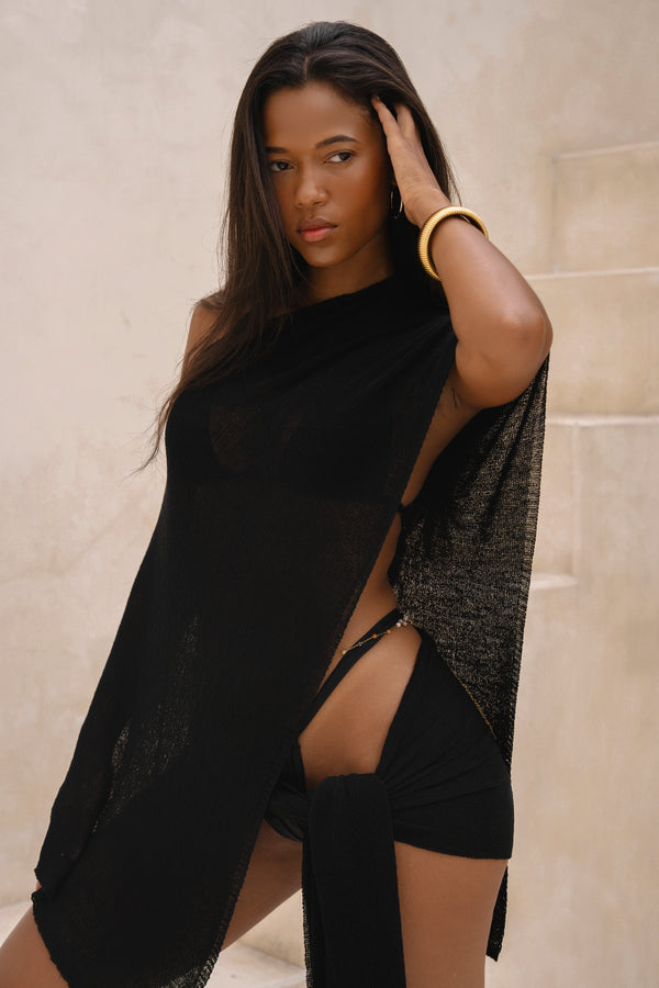 Model wearing a black sarong and black matching cape, posing in front of a neutral background