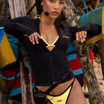 Model on a beach wearing a black long-sleeve zip-up Rogue Season swim top and yellow bikini bottoms with black trim,  with boats in the background