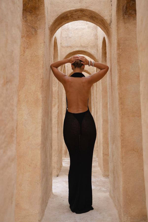 Model wearing a black halter neck dress standing in a sunlit archway with sand-coloured walls.