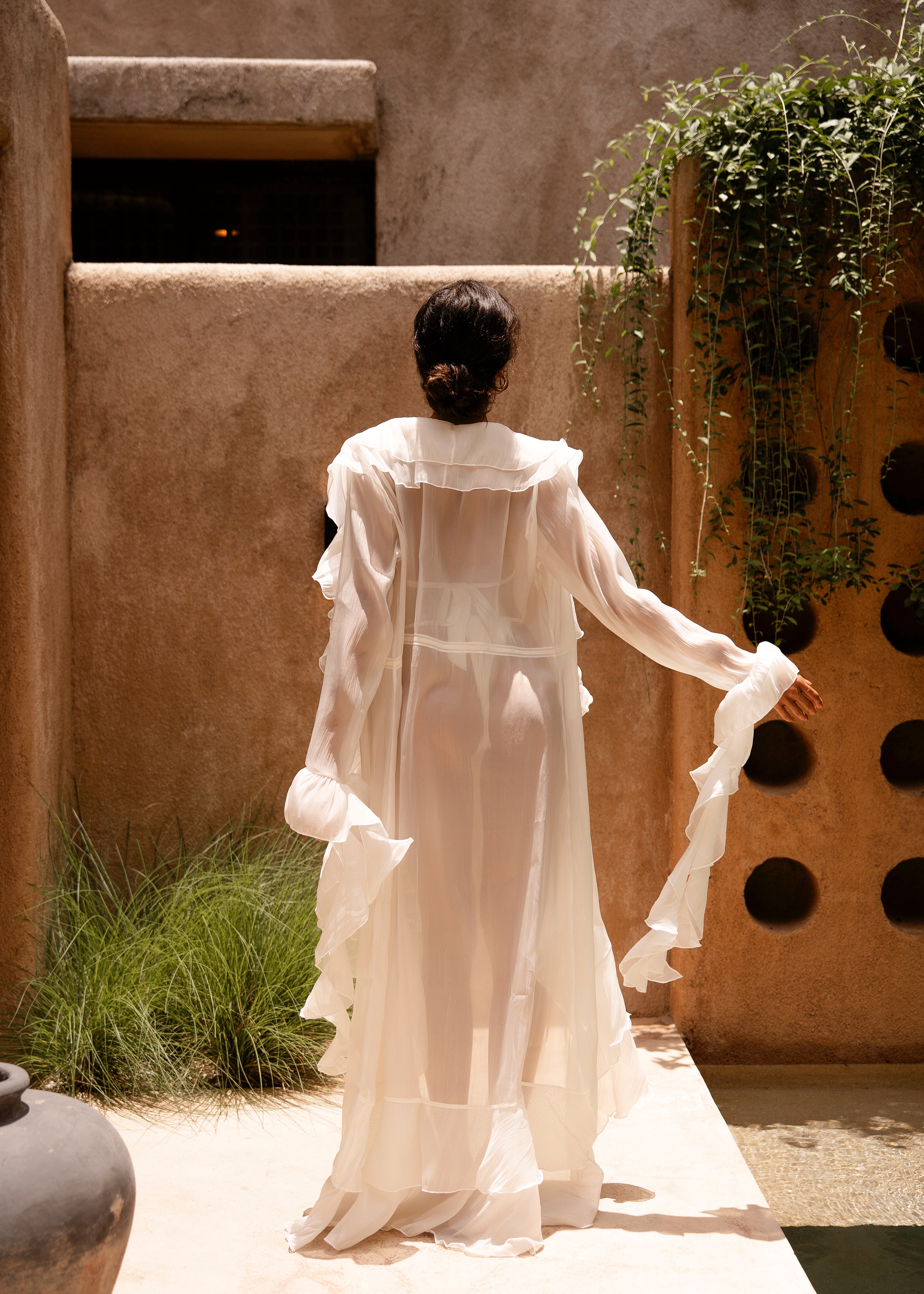 Back shot of model wearing a ivory bikini with a sheer ruffled maxi cover-up, standing outdoors in sunlight against a textured beige wall with greenery.