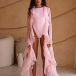 Model wearing a pink swimsuit with a matching sheer robe against a beige wall.