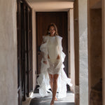 Model wearing a fitted ivory sleeveless bodysuit paired with a draped sheer ivory mini skirt and matching sheer robe walking through a neutral-toned hallway.