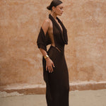 Model wearing a chocolate brown sheer maxi dress with a draped halter neckline, standing side-on against a textured neutral wall.