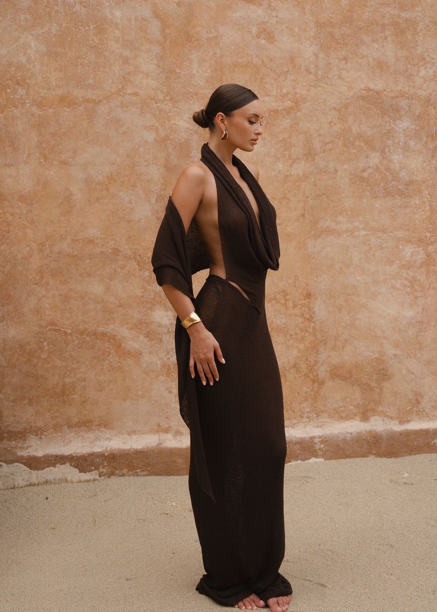Model wearing a chocolate brown sheer maxi dress with a draped halter neckline, standing side-on against a textured neutral wall.