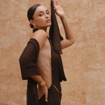 Side image of model wearing a brown halter neck maxi dress standing in a sunlit setting with sand-coloured walls.