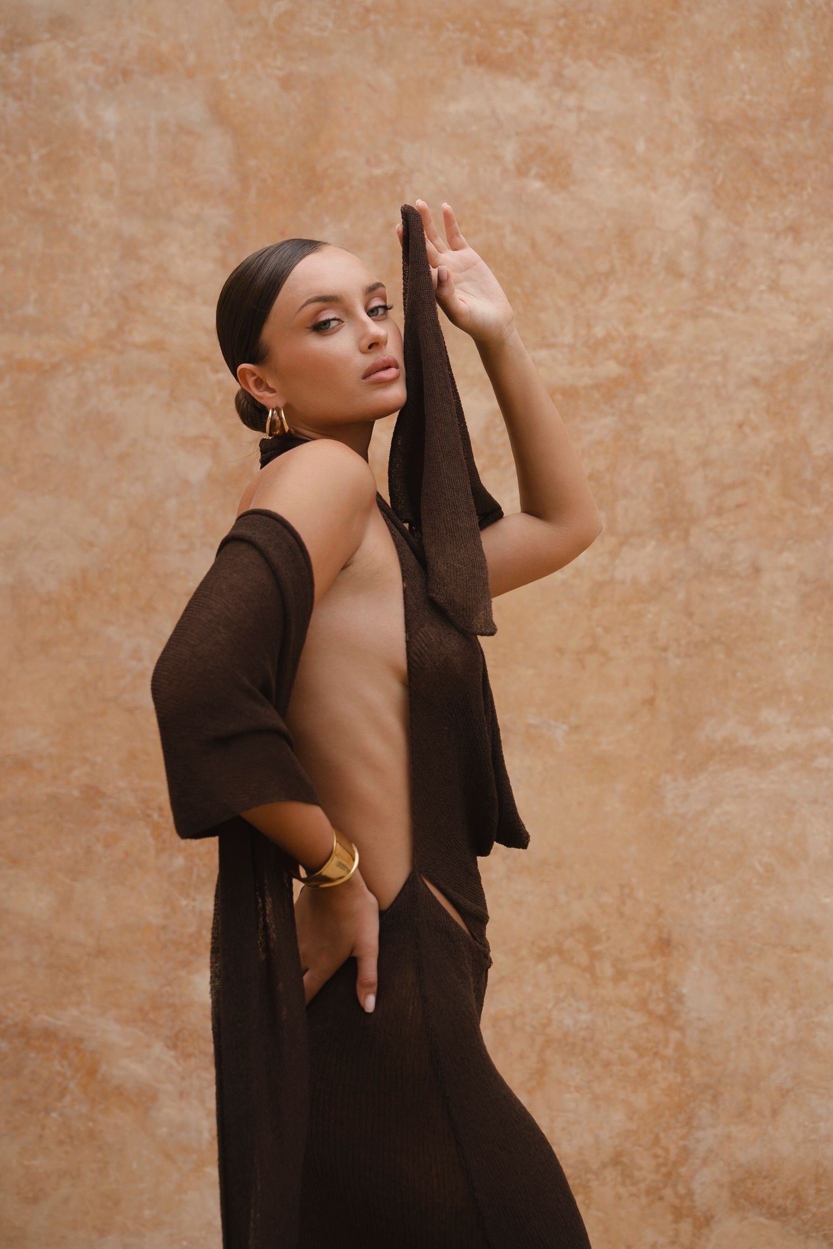 Side image of model wearing a brown halter neck maxi dress standing in a sunlit setting with sand-coloured walls.