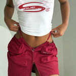 Crop in shot of model wearing a white tank top with a red 'RogueSeason' logo, checkered bikini in red and pink and a red matching skirt.