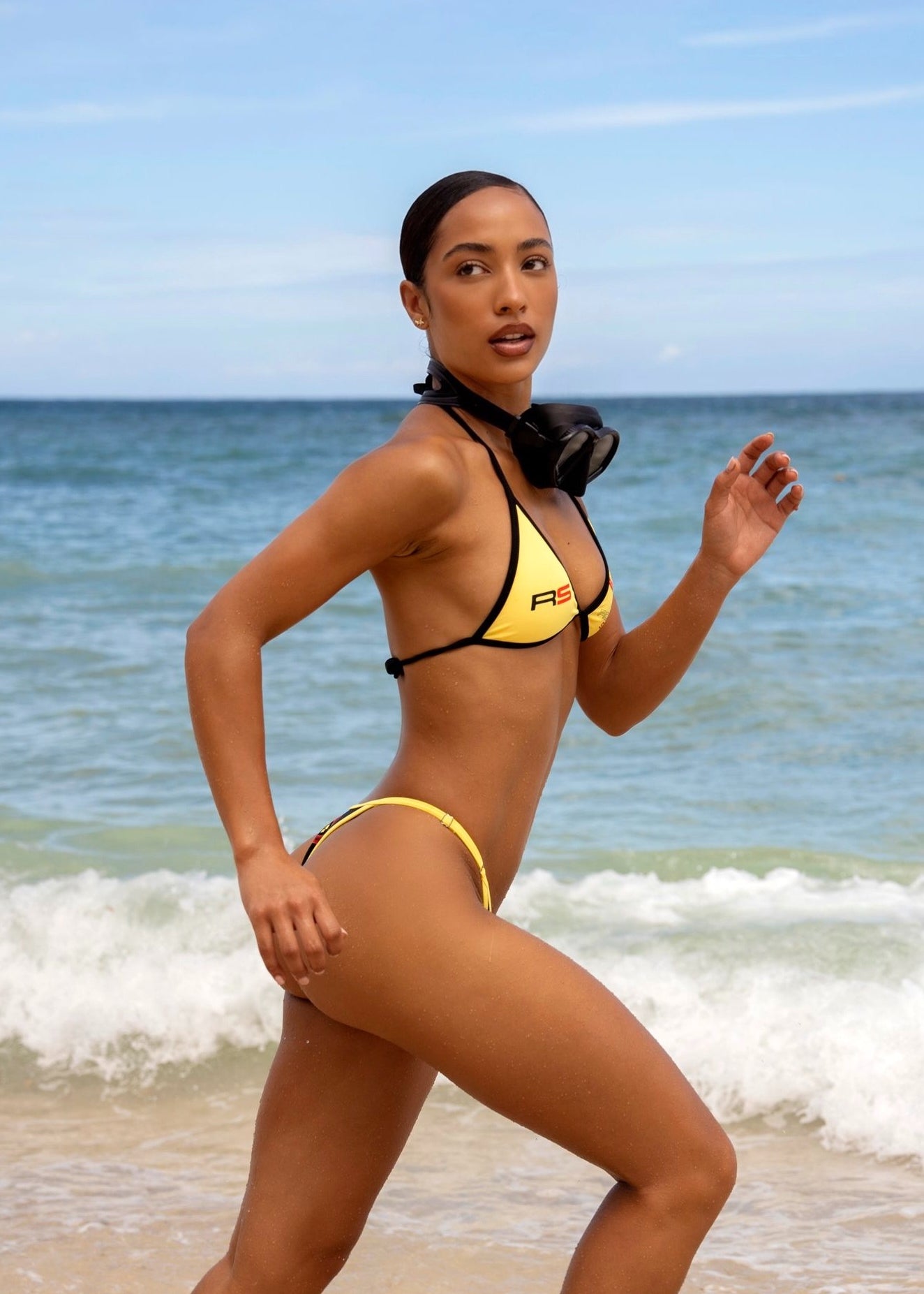 Model in Rogue Season yellow and black bikini running on a beach with ocean waves in the background