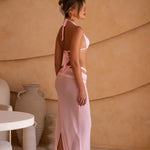 Side shot of model wearing a soft blush pink triangle bikini with adjustable halter straps and minimal-coverage bottoms with a matching sheer maxi skirt. Photographed outdoors, the image highlights the bikini's clean lines, flattering fit and elevated resort-inspired design.