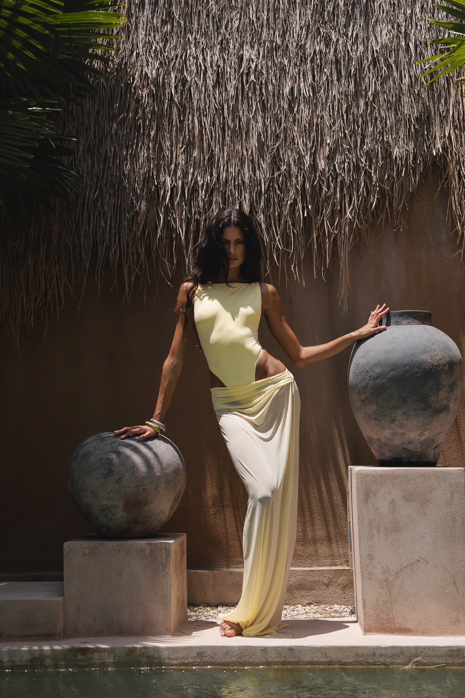 Model in a yellow swimsuit with a matching sheer maxi skirt, standing between two large pots in a tropical setting